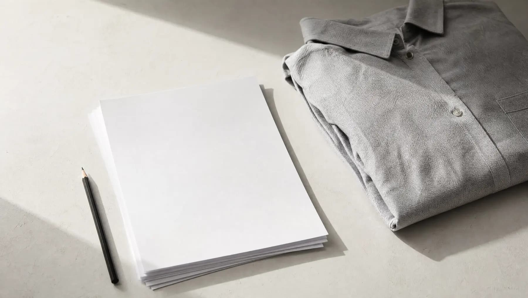Blank printed sheets and a pencil beside a folded shirt on a pale table.